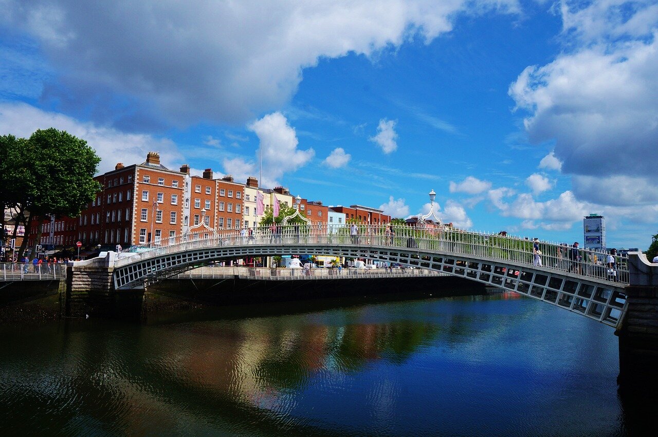 A pedestrian bridge arching over a calm river with a backdrop of colorful buildings under a partly cloudy sky. This bridge is the Ha'penny Bridge, located in Dublin, Ireland.